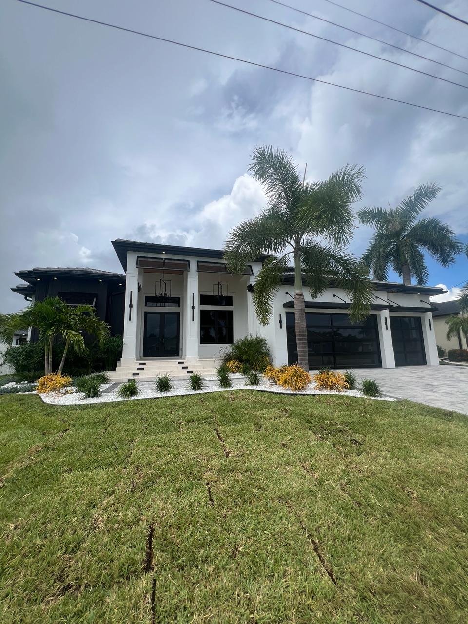 Full facade of a white modern South Florida home with entry and two dark full-view impact glass garage doors framed by palms