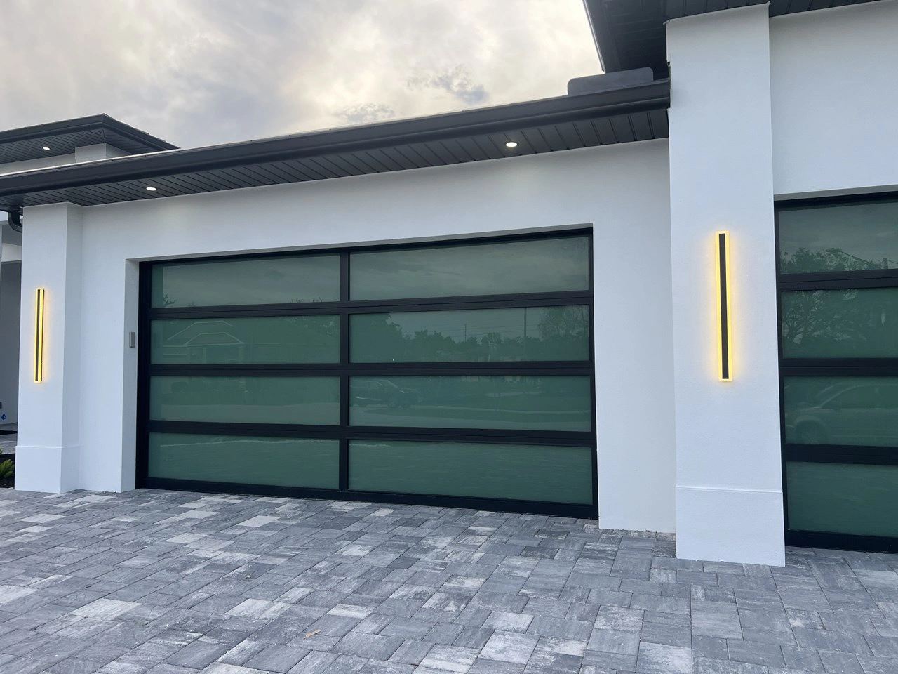 Wide full-view impact glass garage door with green glass and warm accent lighting, palm-frond reflections at dusk